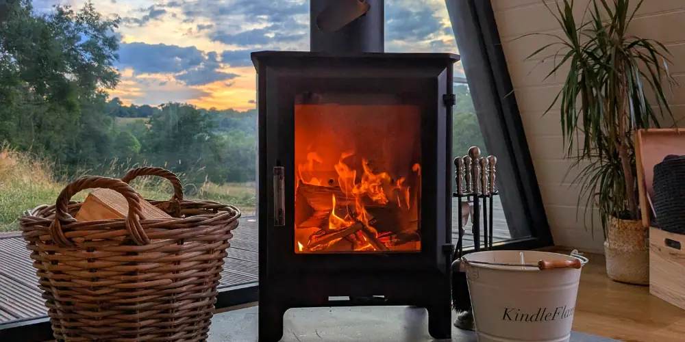 A log basket beside a glowing wood burner at sunset