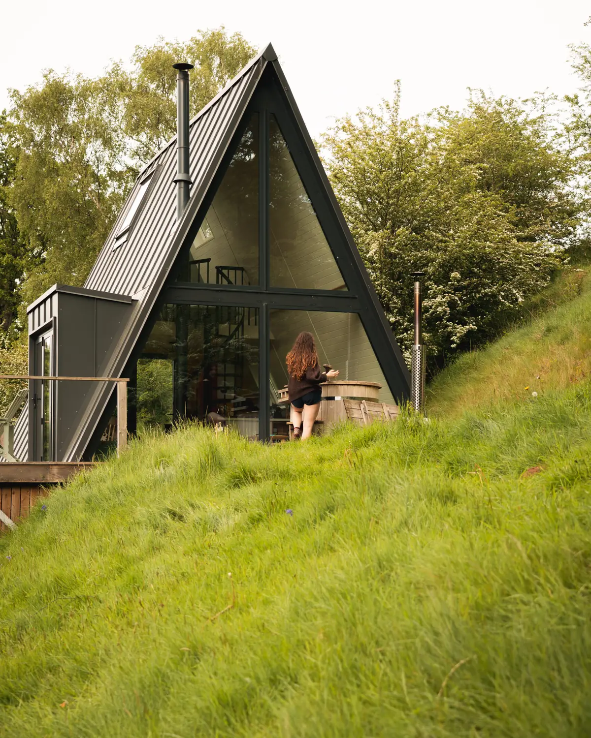 The black A Frame cabin on a grassy slope with trees behind